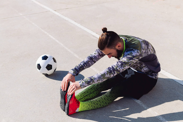 fashionable sportsman stretching legs sitting on the floor next to his football ball in a concrete soccer court, concept of healthy lifestyle and urban sport in the city, copy space for text - Photo, Image