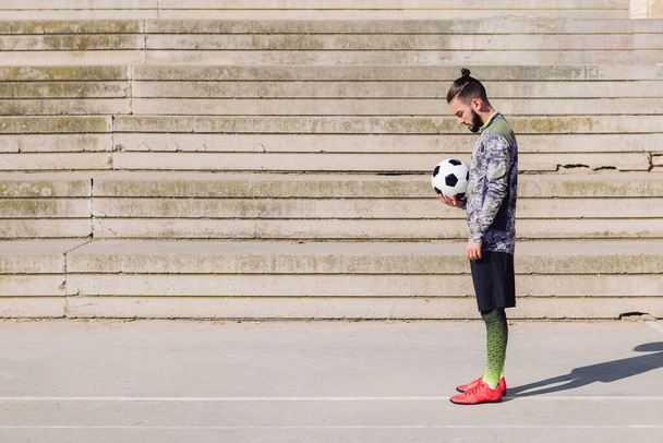 handsome sportsman with hair in a bun standing with the ball in his hand on a concrete soccer court, concept of healthy lifestyle and urban sport in the city, copy space for text - Photo, Image