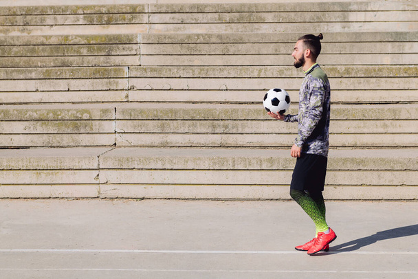 handsome sportsman with hair in a bun walking with the ball in his hand on a concrete soccer court, concept of healthy lifestyle and urban sport in the city, copy space for text - Photo, Image