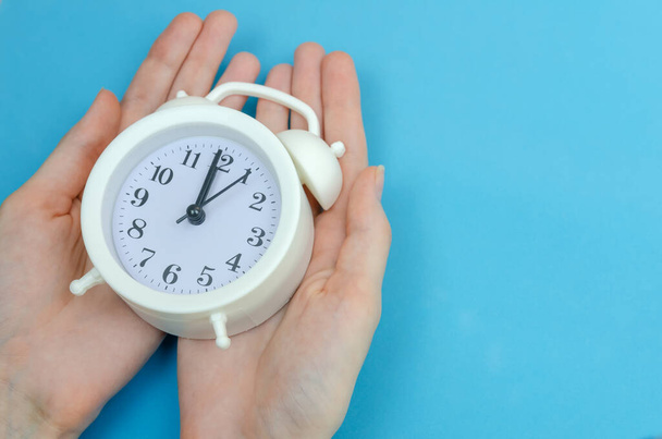 Las manos femeninas sostienen un reloj despertador blanco sobre un fondo azul, espacio para copiar. - Foto, Imagen