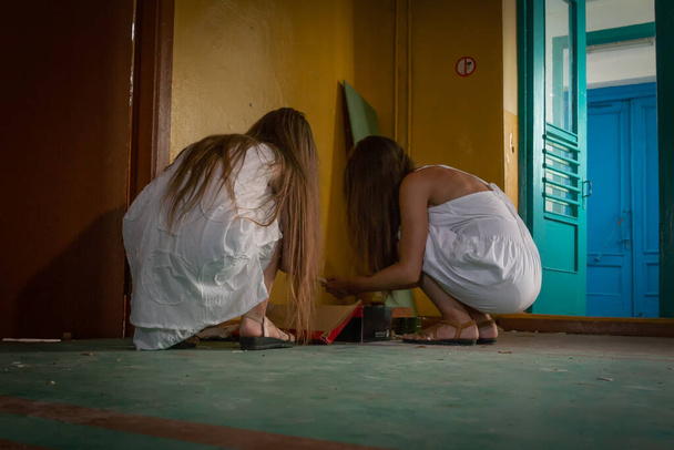 Duas meninas em vestidos brancos com cabelos longos estão sentadas no chão no salão de um prédio abandonado e estão classificando através da mukalutura, uma foto da parte de trás. Conceito de horror, misticismo - Foto, Imagem