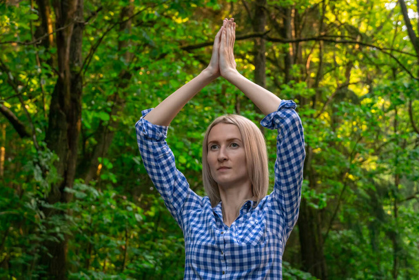 young beautiful blonde woman holding her hands above her head is playing sports in the forest at sunset. concept of yoga. - Фото, изображение