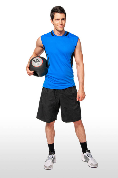Portret van een fitte jonge blanke mannelijke atleet met kort donker haar die in zijn eentje poseert en een zwarte medicijnbal vasthoudt in een studio met een witte achtergrond in een zwarte broek en een blauw mouwloos shirt. - Foto, afbeelding