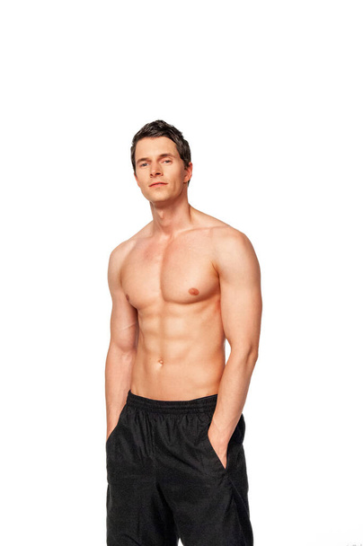 Portrait of a confident young white male athlete with short dark hair posing by himself with hands in his black short pockets in a studio with a white background. - Фото, зображення