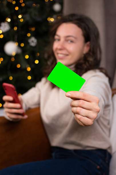 Bulanık gülümseyen kadın, boş kredi kartının önünde kameraya bakıyor, akıllı telefon kullanıyor ve evde Noel ağacının yanında oturuyor. İnternetten yeni yıl alışverişi. Dikey - Fotoğraf, Görsel