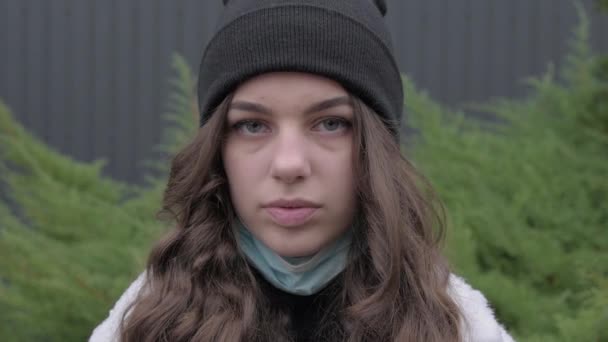 Looks serious young adult european woman in medical mask pretty face looking at camera posing alone standing at outdoor. front portrait. In warm clothes, jacket and hat in the yard. - Filmagem, Vídeo