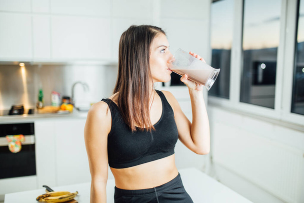 Woman in sportswear drinking sweet banana chocolate protein powder milkshake smoothie.Drinking protein after at home workout.Whey, banana and low fat milk sports nutrition diet.Healthy lifestyle - Foto, afbeelding