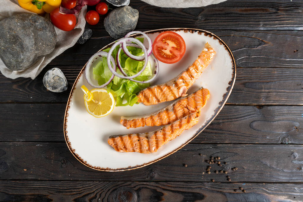 Lachssteak vom Grill mit Salat, Zwiebeln und Tomaten. Klassisches warmes Fischgericht mit Meeresfisch, Gemüse und Zitronensalat in einem Keramikteller auf einem hölzernen Küchentisch. - Foto, Bild