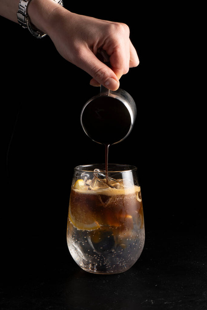 Girl bartender prepares a cocktail and pours coffee into an espresso tonic. Cold alcoholic cocktail of coffee and a refreshing tonic in a transparent glass on a black background. - Foto, Bild