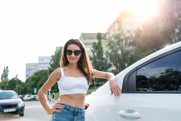 woman in the summer stands by the car, a white sedan, a beautiful girl smiles, long hair sunglasses. Denim shorts and a white top. - Φωτογραφία, εικόνα