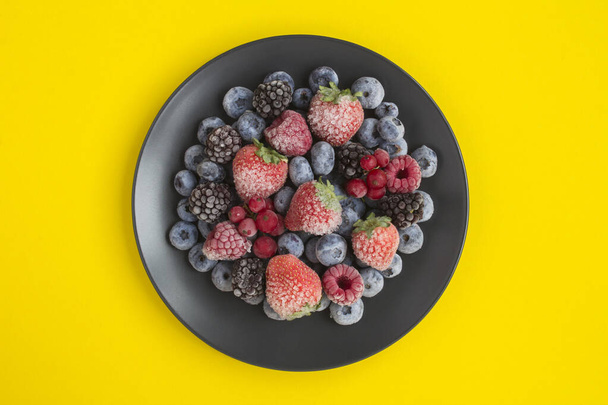 Top view of frozen berries on the black  plate in the center of the yellow background - Photo, Image