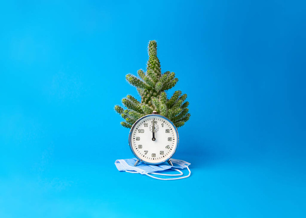 Die alte blaue Analoguhr, die genau Mitternacht anzeigt, trägt eine medizinische Schutzmaske, und dahinter steht ein Kaktus, der aussieht wie ein Weihnachtsbaum. Dahinter ist ein blauer Hintergrund. - Foto, Bild