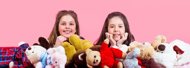 Little girls play with teddy bears on a pink background. Two beautiful happy girls are lying and embracing plushs toy in children room. Girl hugging teddybears, childhood - Zdjęcie, obraz