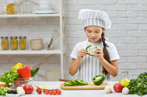 маленькая девочка в шляпе шеф-повара и фартуке, режущая зеленый перец - Фото, изображение