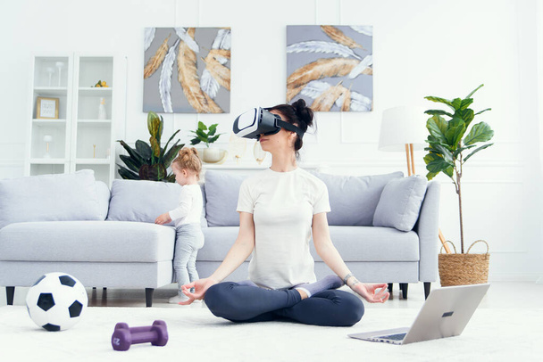 Young mom meditating in lotus yoga position using vr goggles while her children playing at home on background. - Foto, immagini