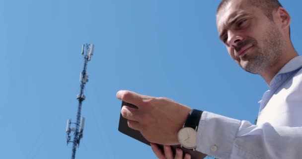 Een man verbindt zijn tablet met het 5G-netwerk terwijl hij op de achtergrond van een communicatietoren staat. - Video