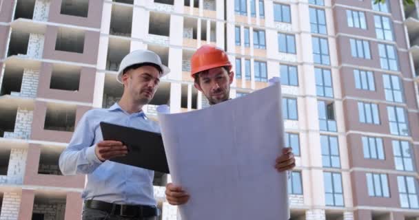 Un ingénieur avec une tablette explique et montre au constructeur le papier et les dessins électroniques, puis pointe vers un bâtiment résidentiel en construction. - Séquence, vidéo