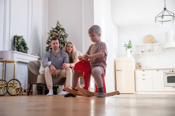El pequeño hijo monta un caballo de madera mientras los padres jóvenes se relajan en casa en el sofá, el niño se divierte, el ocio familiar en la sala de estar, la familia amigable y feliz - Foto, Imagen