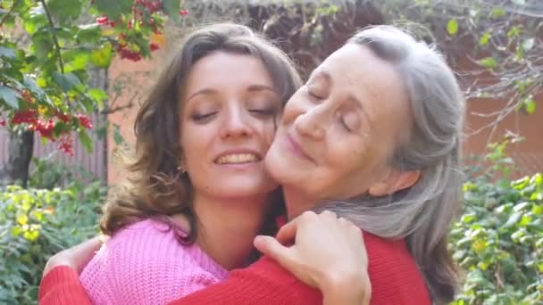 Senior mother with gray hair with her adult daughter looking at the camera in the garden and hugging each other during sunny day outdoors - Footage, Video