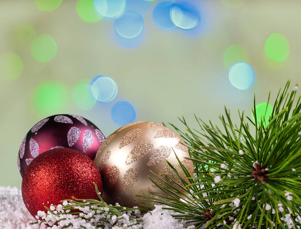 Een groep van drie kerstballen en een dennentak in de sneeuw. De achtergrond is groenachtig bokeh met blauwe en licht groene lichten.De ballen zijn bordeaux, geel en rood.Kopieer ruimte. - Foto, afbeelding
