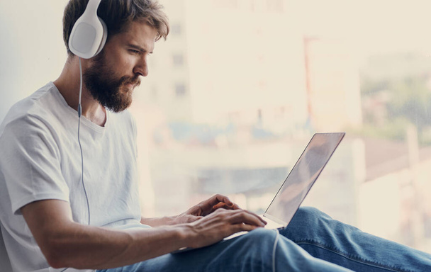 άνθρωπος με φορητό υπολογιστή κάθεται στο παράθυρο πληκτρολόγιο οθόνη ακουστικά μοντέλο - Φωτογραφία, εικόνα