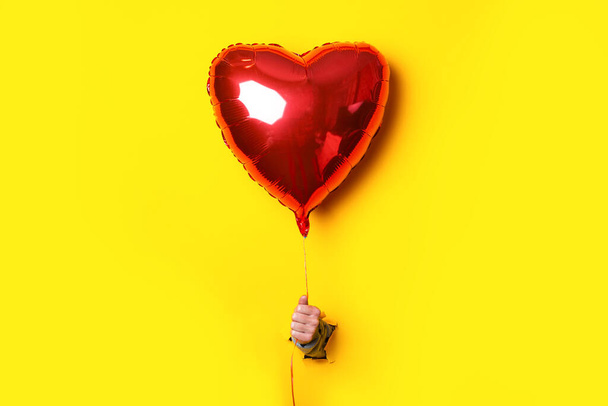 Žena ruka v roztržené žluté pozadí drží vzduch nafukovací červené srdce balón - Fotografie, Obrázek