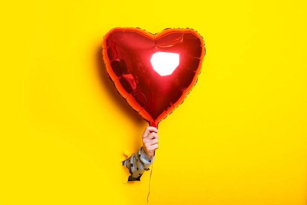 Female hand in a torn yellow background holding an air inflatable red balloon heart - Foto, Imagem