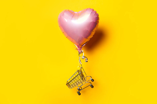 prázdný nákupní vozík mouchy na růžové srdce ve tvaru vzduchového balónu na žlutém pozadí - Fotografie, Obrázek