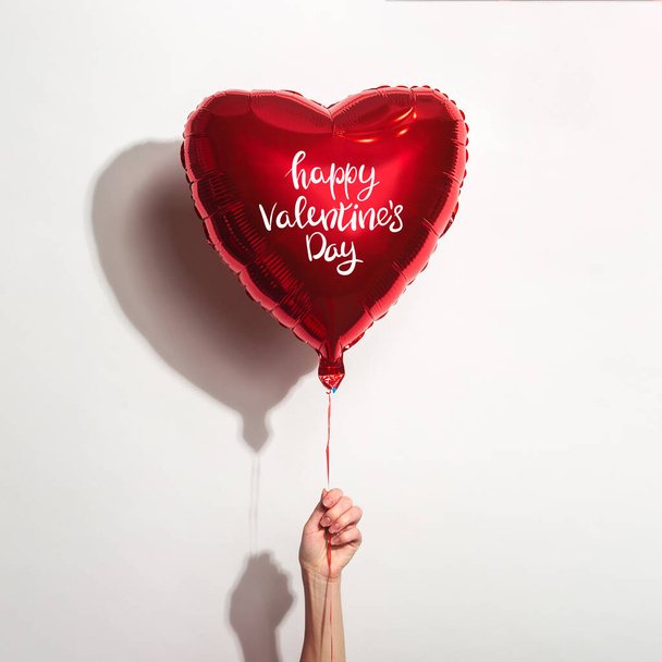 mano femenina sostiene un globo de aire en forma de corazón con la inscripción Feliz Día de San Valentín sobre un fondo blanco. - Foto, Imagen
