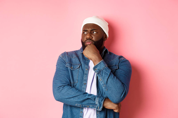Modelo masculino afroamericano reflexivo serio que toma la decisión, mirando a la izquierda preocupada, pensando o teniendo dudas, de pie sobre fondo rosado - Foto, imagen