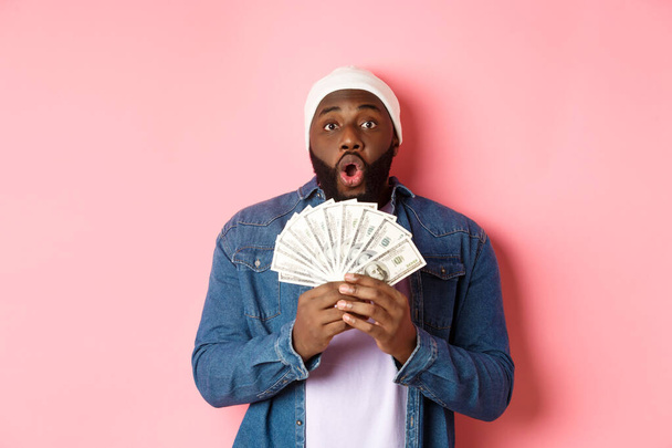 Homem afro-americano incrível recebe prêmio em dinheiro, mostrando dinheiro e olhando para a câmera com espanto, de pé sobre fundo rosa - Foto, Imagem