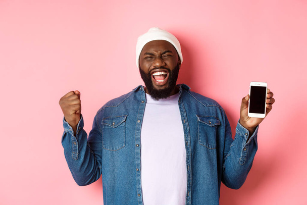 Chico negro feliz celebrando el logro en línea, mostrando la pantalla del teléfono inteligente, regocijo y gritar sí con satisfacción, de pie sobre el fondo rosa - Foto, Imagen