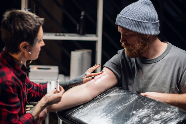 Jovem artista de tatuagem de menina informal coloca uma bandagem com vaselina na mão de um jovem depois de aplicar com sucesso uma tatuagem em sua mão. - Foto, Imagem