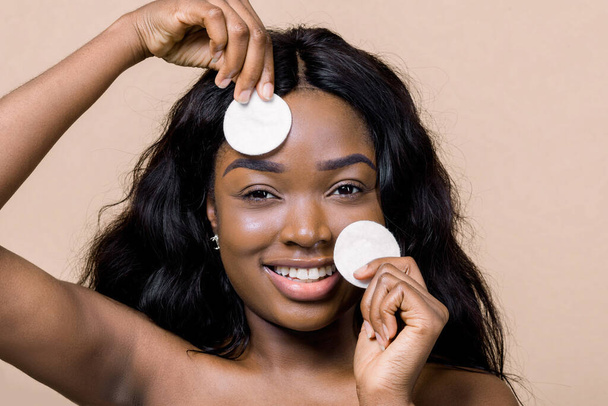 Young pretty african woman posing to camera with two cotton wipe sponges, taking off makeup, on isolated beige background. Cleansing face, daily healthy beauty routine, skincare concept - Valokuva, kuva