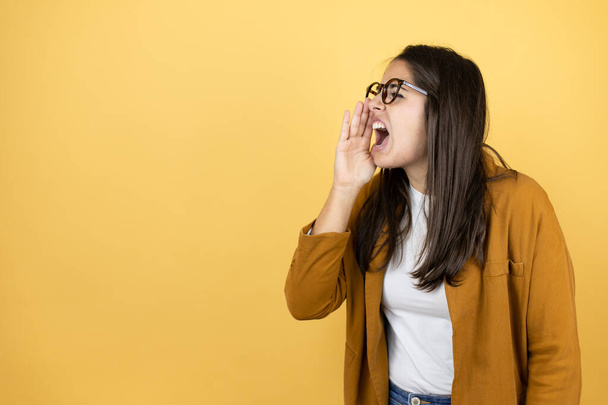 Jonge mooie vrouw draagt een blazer over geïsoleerde gele achtergrond schreeuwen en schreeuwen luid naar zij met handen op de mond - Foto, afbeelding