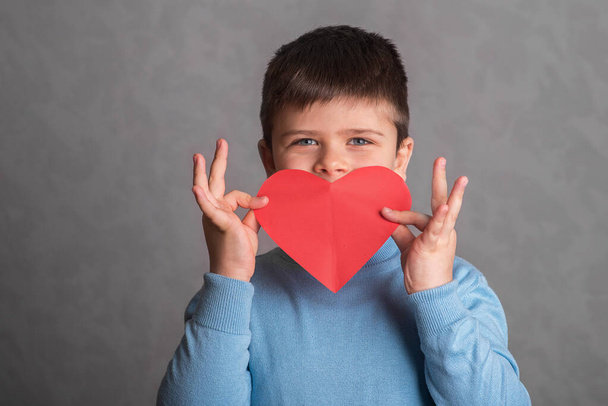 A gyermek egy papírból kivágott nagy vörös szív mögé rejtette arcát. A fiú vörös papírból kivágott szívet mutat. A gyermek kezében egy piros papírból készült valentin szív formájában. A szürke hátterű baba a szerelem szimbólumát mutatja. Egy boldog gyerekműsor - Fotó, kép