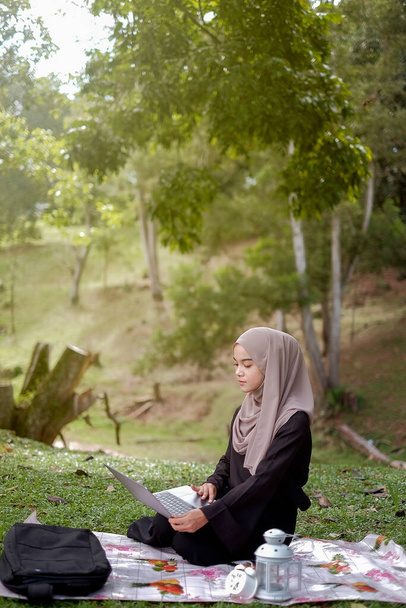 Фото 20-летней студентки-исламистки в платке, сидящей в зеленом парке и использующей серебряный ноутбук. - Фото, изображение