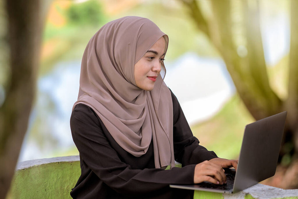 Foto de una hermosa estudiante islámica de 20 años con pañuelo en la cabeza sentada en el parque verde y usando una computadora portátil de plata. - Foto, imagen