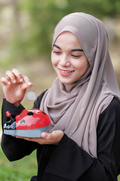 Piękna młoda kobieta z hidżabem wkładająca monety do skarbonki w parku. Koncepcja oszczędzania pieniędzy. - Zdjęcie, obraz