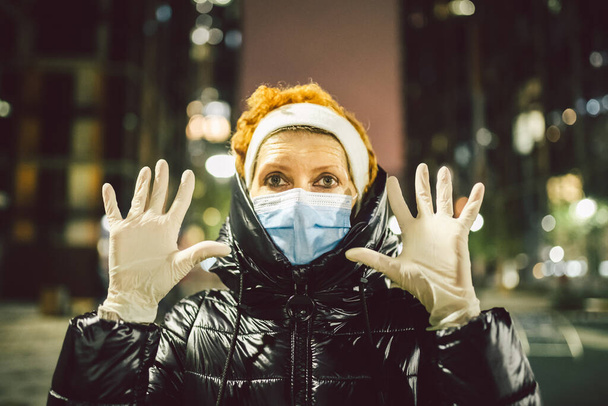 Λευκά ιατρικά γάντια φόρεμα στα χέρια ενήλικη γυναίκα στο δρόμο της πόλης Ευρώπη κατά τη διάρκεια της καραντίνας κλειδώματος covid19, προστατευτικό πρόσωπο μάσκα, cityview βράδυ.. - Φωτογραφία, εικόνα