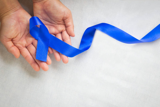 Hands holding deep blue ribbon on white fabric with copy space. Colorectal Cancer Awareness, Colon cancer of older person and world diabetes day, Child abuse prevention. Healthcare, insurance concept. - Fotoğraf, Görsel