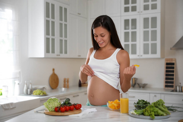 Young pregnant woman with slice of bell pepper near table in kitchen. Taking care of baby health - Photo, image