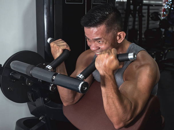 Un macho asiático buff hace algunos rizos de máquina predicador en el gimnasio. Entrenamiento de brazos y bíceps. Fitness en hombres de 40 años - Foto, Imagen