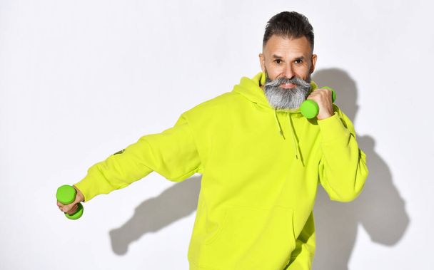 Happy smiling old brutal bearded man in yellow hoodie enjoys fitness training with green dumbbells in hands - Φωτογραφία, εικόνα
