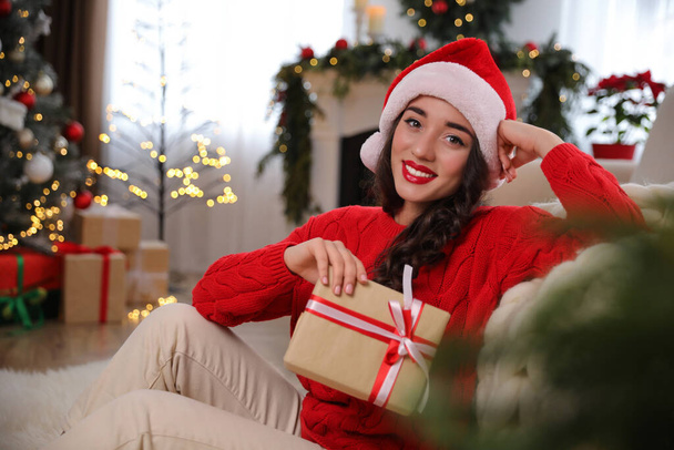 Beautiful woman wearing Santa hat with Christmas gift on floor at home - Fotografie, Obrázek