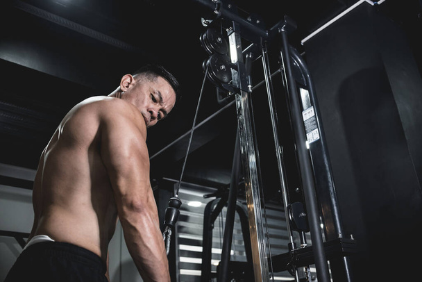 Un hombre asiático buff y sin camisa haciendo ejercicio en el gimnasio, haciendo flexiones de cable. Entrenamiento de entrenamiento de brazo de tríceps - Foto, Imagen