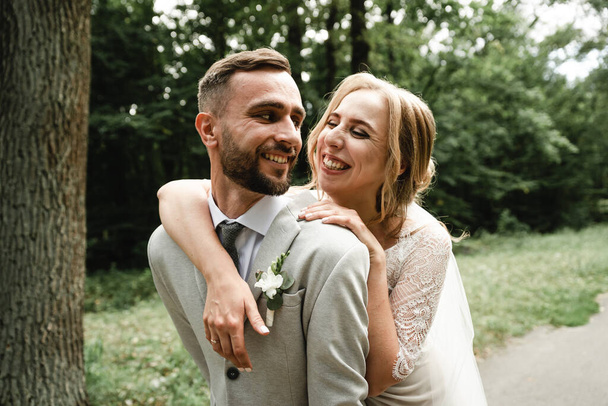 the bride gently hugs the groom from the back and happily whispers in his ear the kind words on the wedding day,Wedding couple. Happy bride with wedding bouquet holds the groom's arm. Stylish newlywed couple. Marriage concept. Rustic wedding - Zdjęcie, obraz