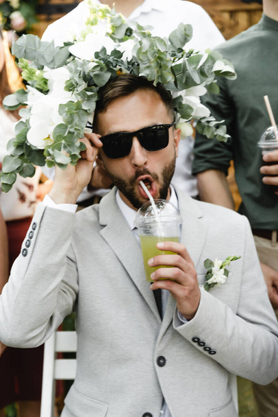 the groom in sunglasses sits on a chair in one hand holds a glass with juice from which he drinks through a straw and on whose head lies a wedding bouquet,happy wedding - Photo, Image