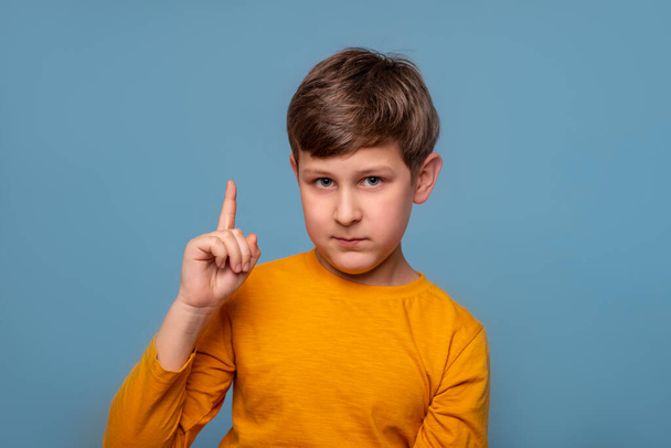 Studio shot van jonge jongen dragen geel shirt bewijzen iets door het opheffen van zijn wijsvinger tegen een blauwe achtergrond met kopieerruimte  - Foto, afbeelding