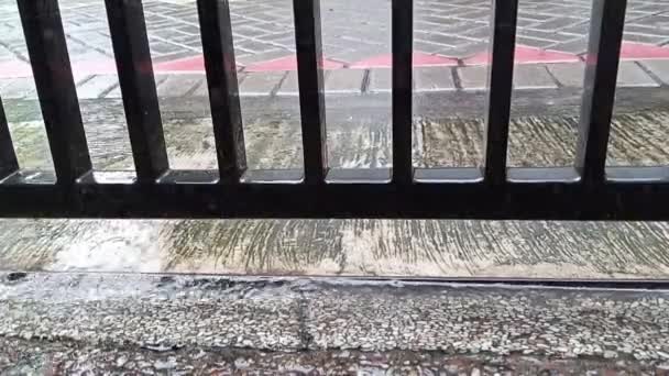 капли дождя падают на бетон и черный железный забор на тротуаре пешеходной улицы в городской застройке - Кадры, видео